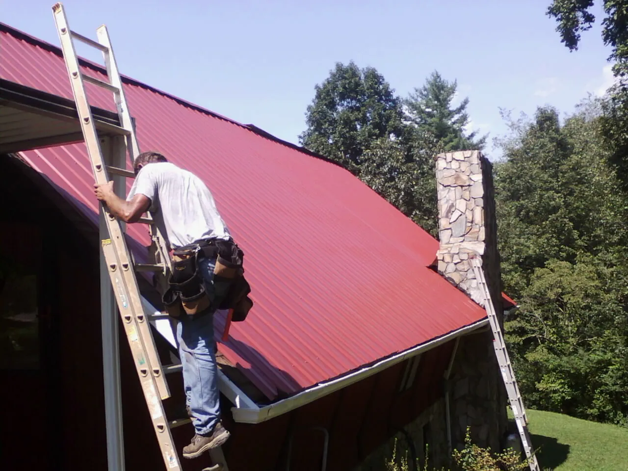 Roofer on a ladder beside a red metal roof and stone chimney — Soffit & Fascia Repair by Roofing Brothers in Clay Center