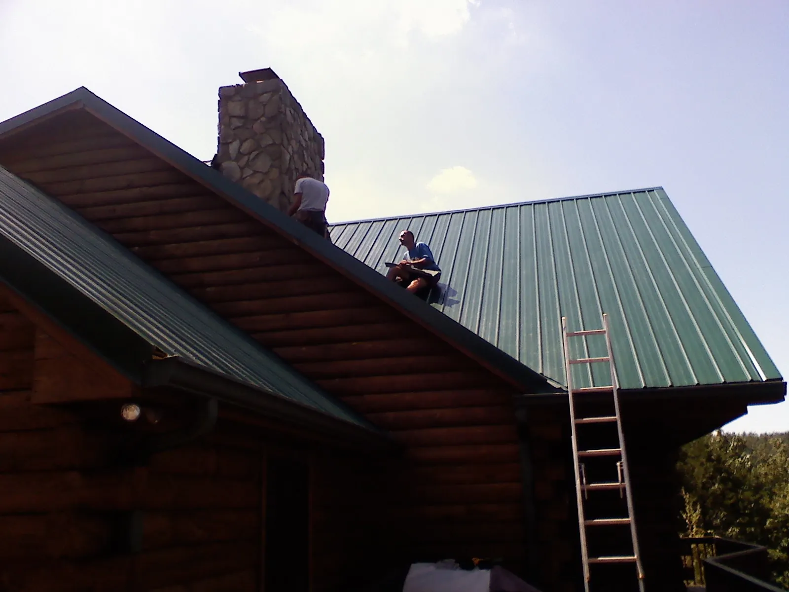Roofers installing green metal panels beside a stone chimney on a log home in Clay Center
